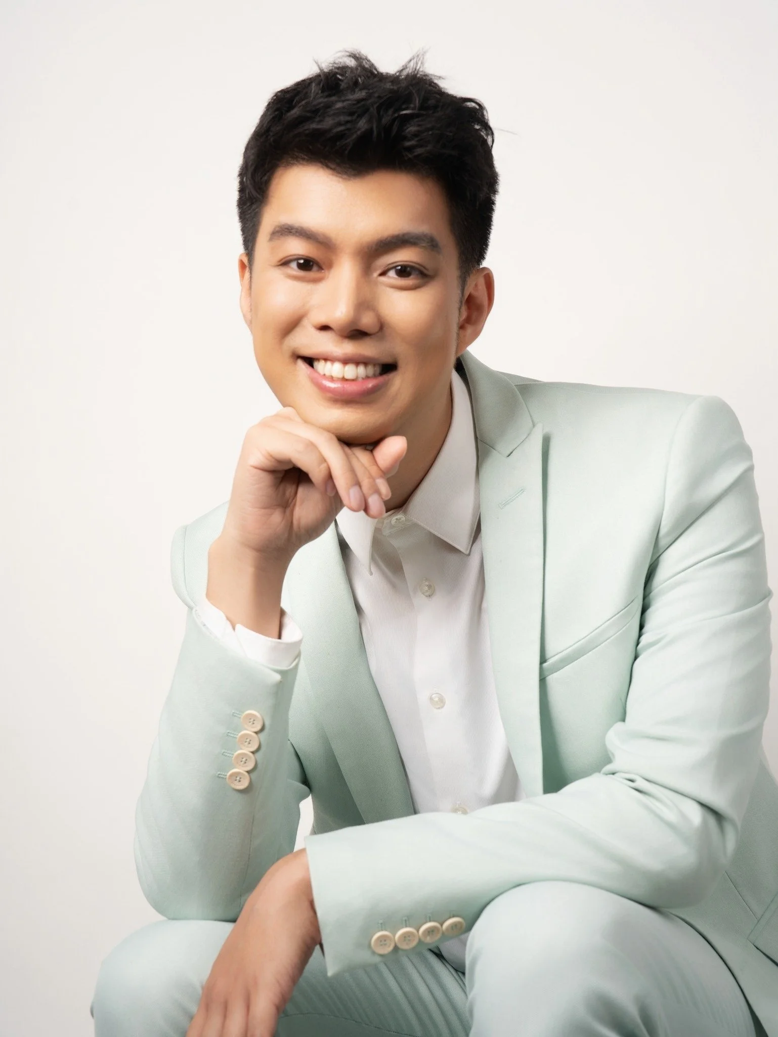 A young man dressed in a light-colored suit, smiling and with his chin resting on his hand, posed against a plain background.