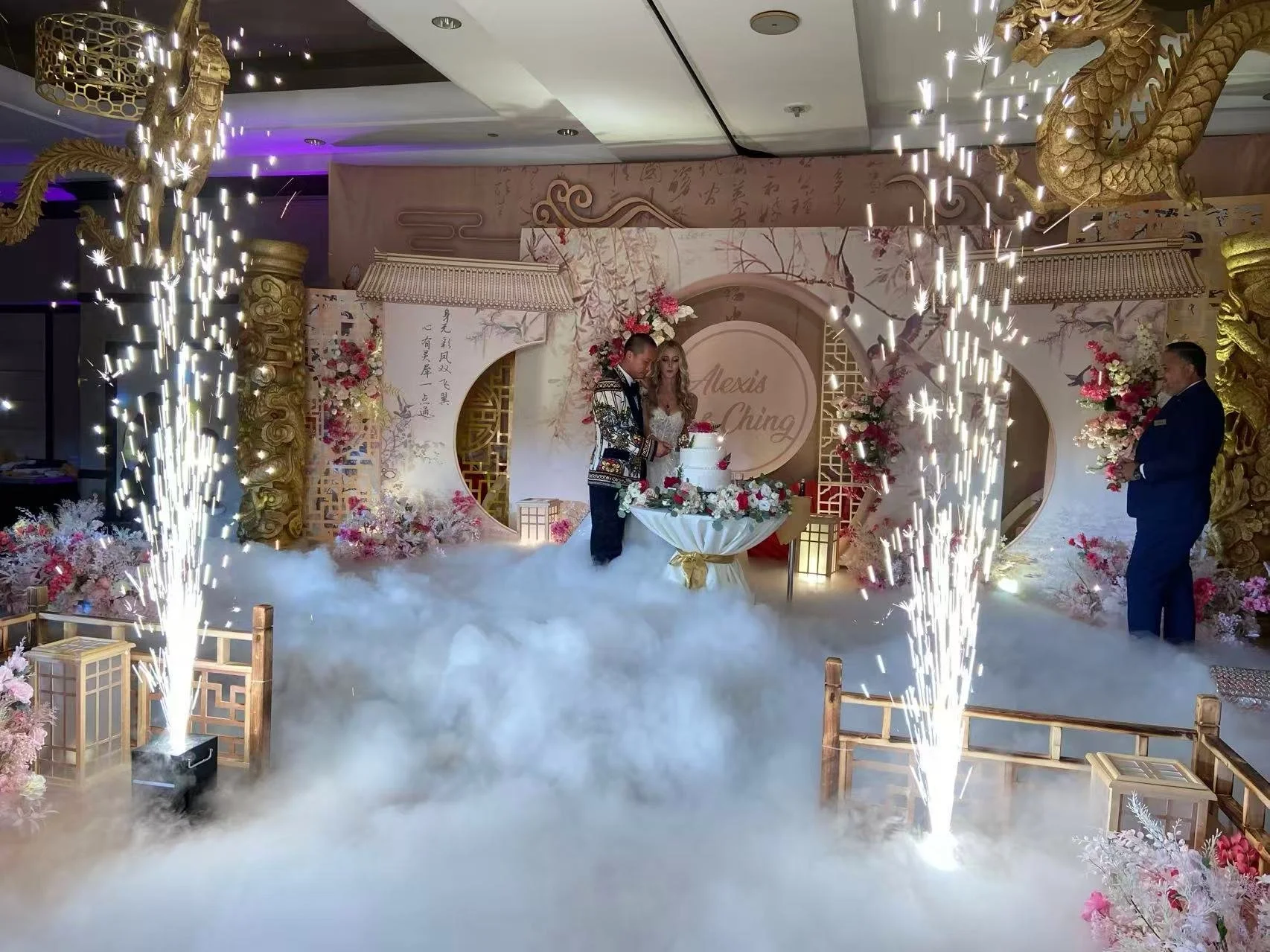 A wedding celebration with a couple cutting a wedding cake on a decorated stage with pink and white flowers, golden dragon decorations, and pyrotechnics, with a fog effects on the floor.