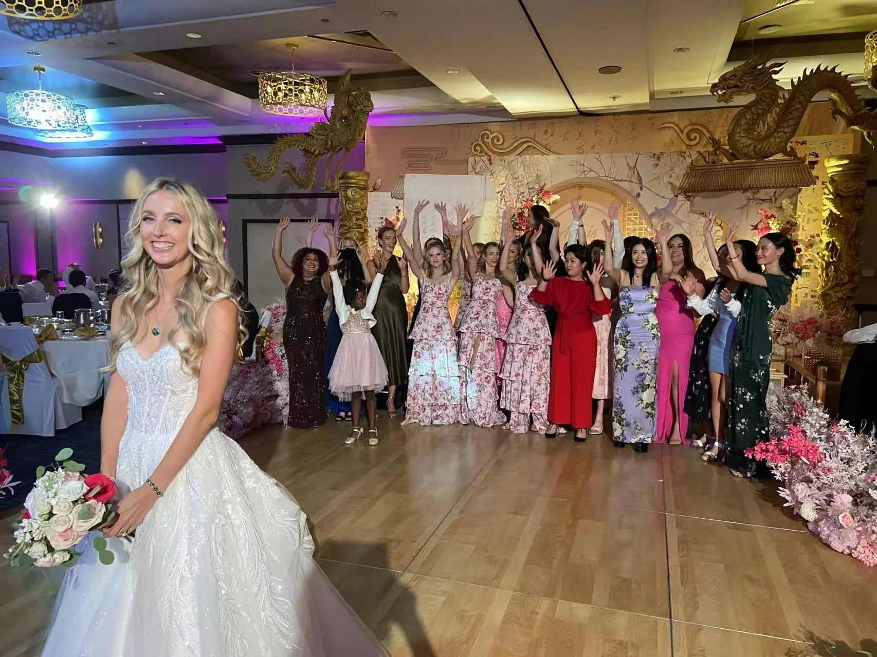 A bride in a white wedding dress holding a bouquet of flowers stands in the foreground, smiling, at a celebration with a group of women in colorful dresses posing and dancing in the background, under decorative lighting and dragon sculptures.