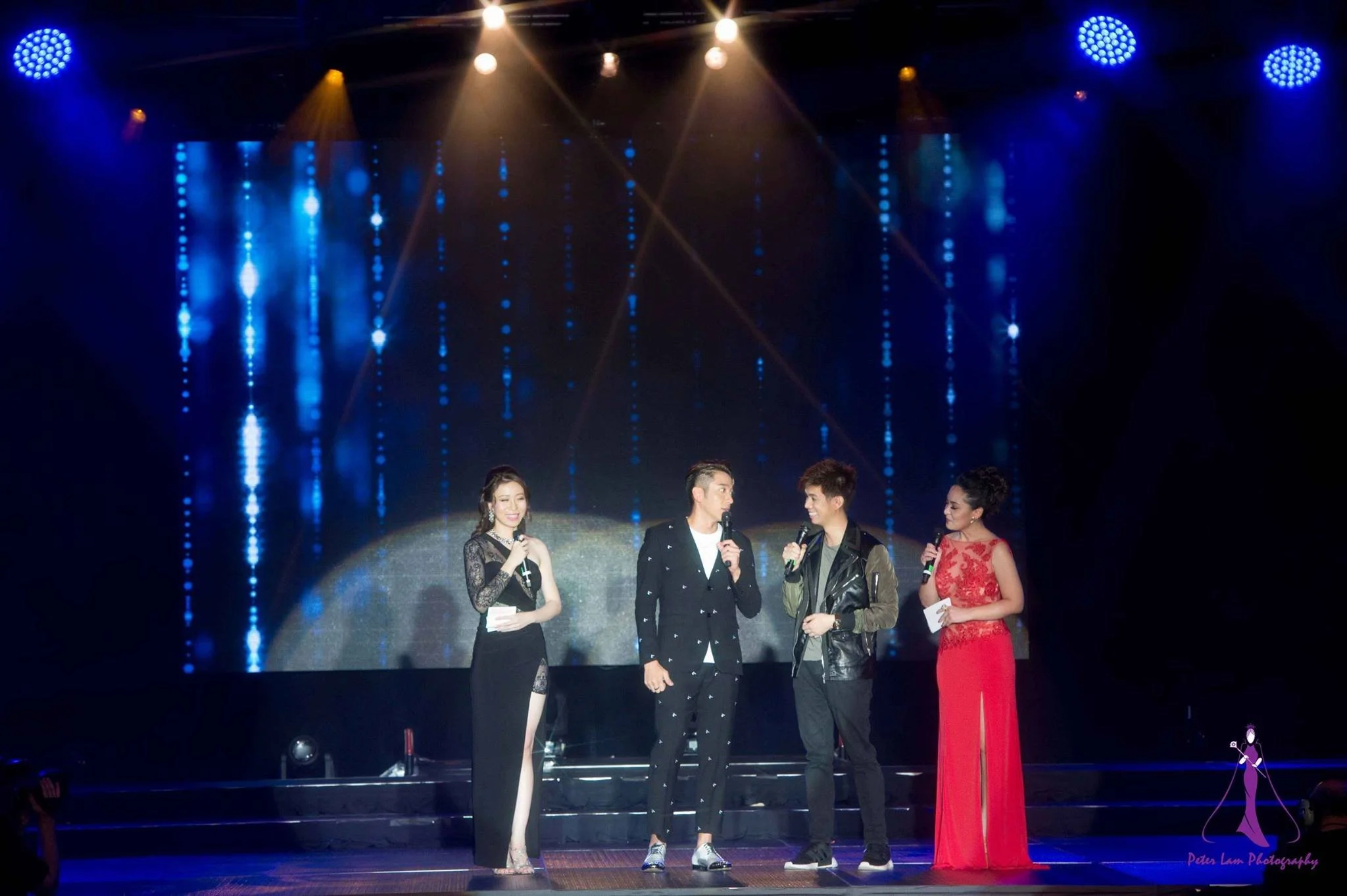 Four performers standing on a stage, three holding microphones, with a DJ or host on the left, a man and woman in the center, and a woman in a red gown on the right. The stage is illuminated with blue and yellow lights, and a large screen with blue a
