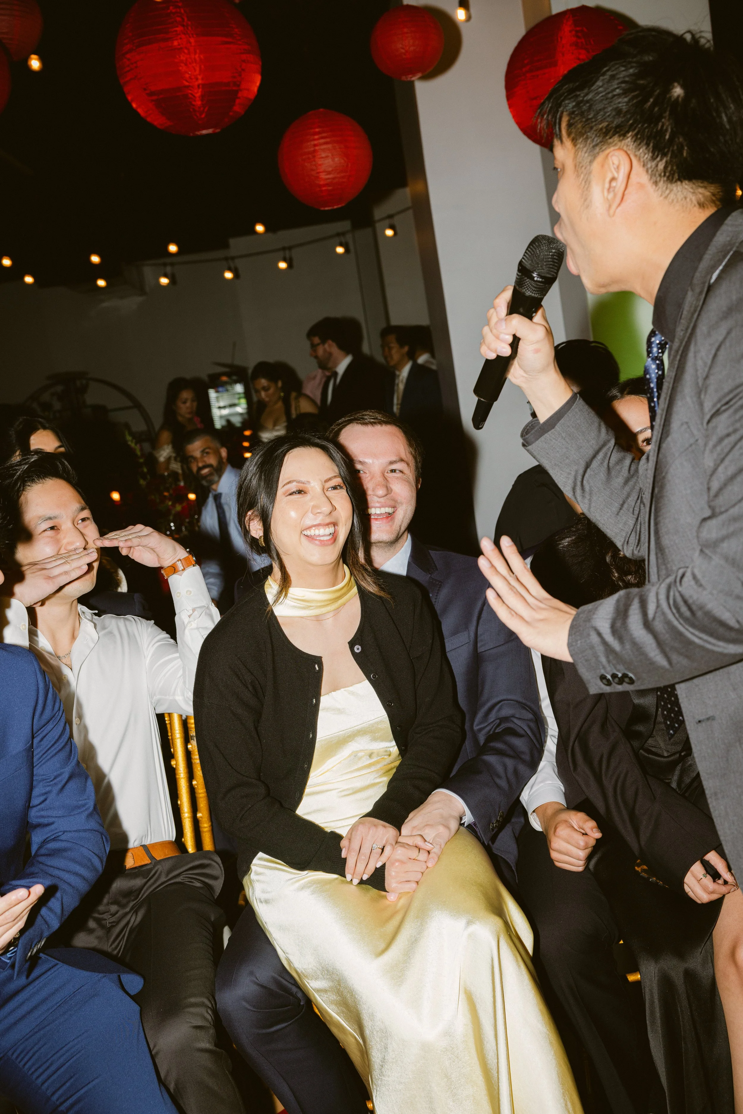 A man with a microphone performing for an audience at a celebration or event, with smiling and laughing guests seated nearby.