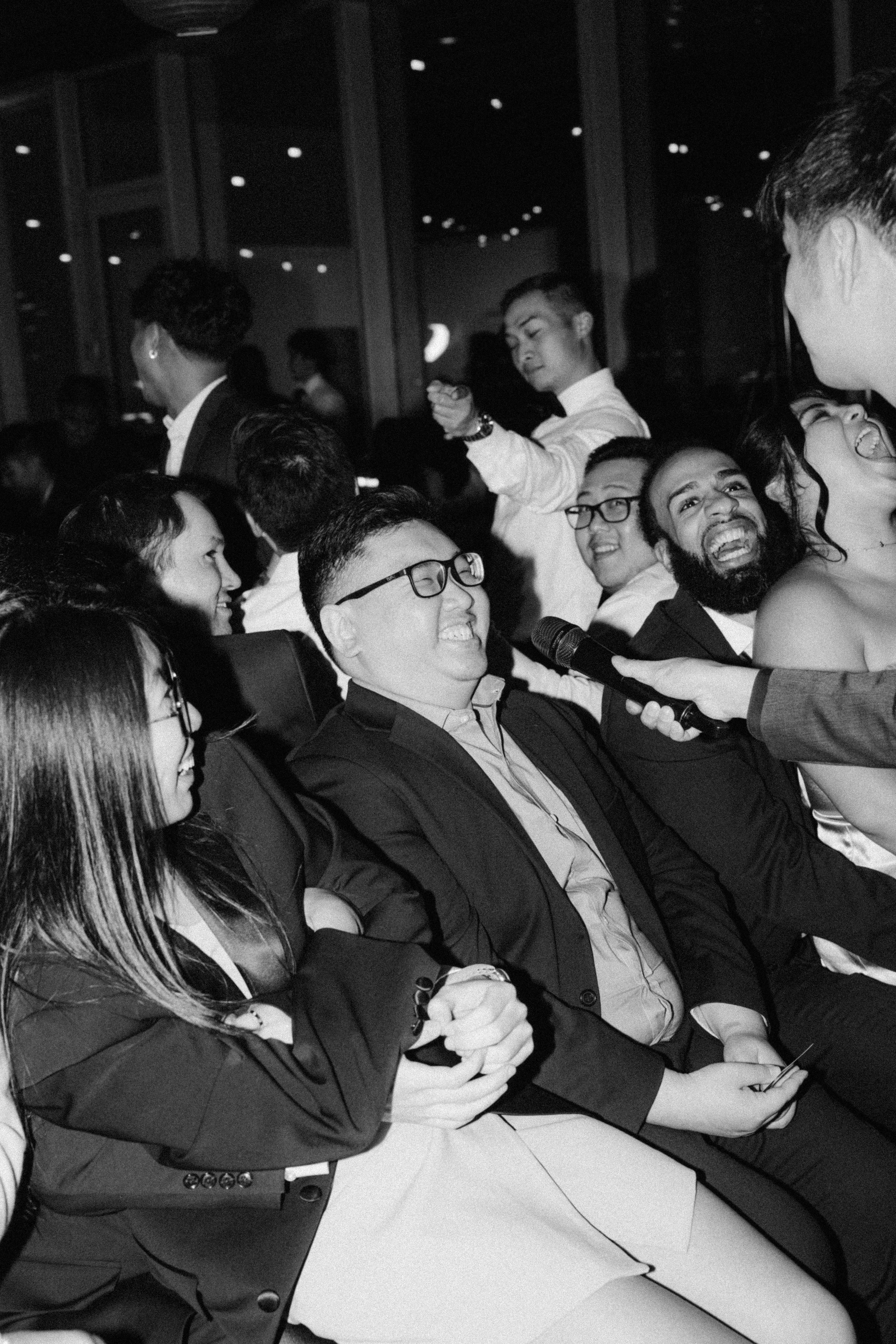 People sitting in an audience, smiling and laughing during an event, with a woman holding a microphone in the foreground.
