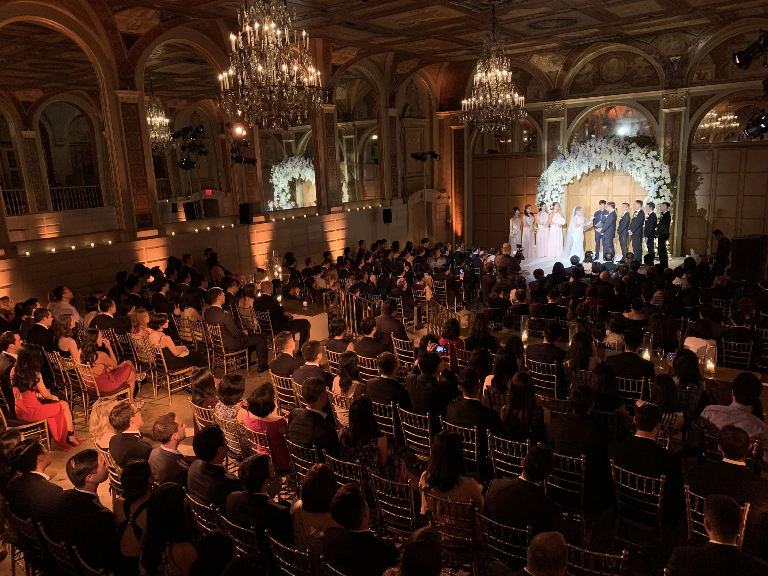 A wedding ceremony with the bride and groom along with bridesmaids and groomsmen on stage under a floral arch, with an audience seated in chairs watching in a grand hall decorated with chandeliers and warm lighting.