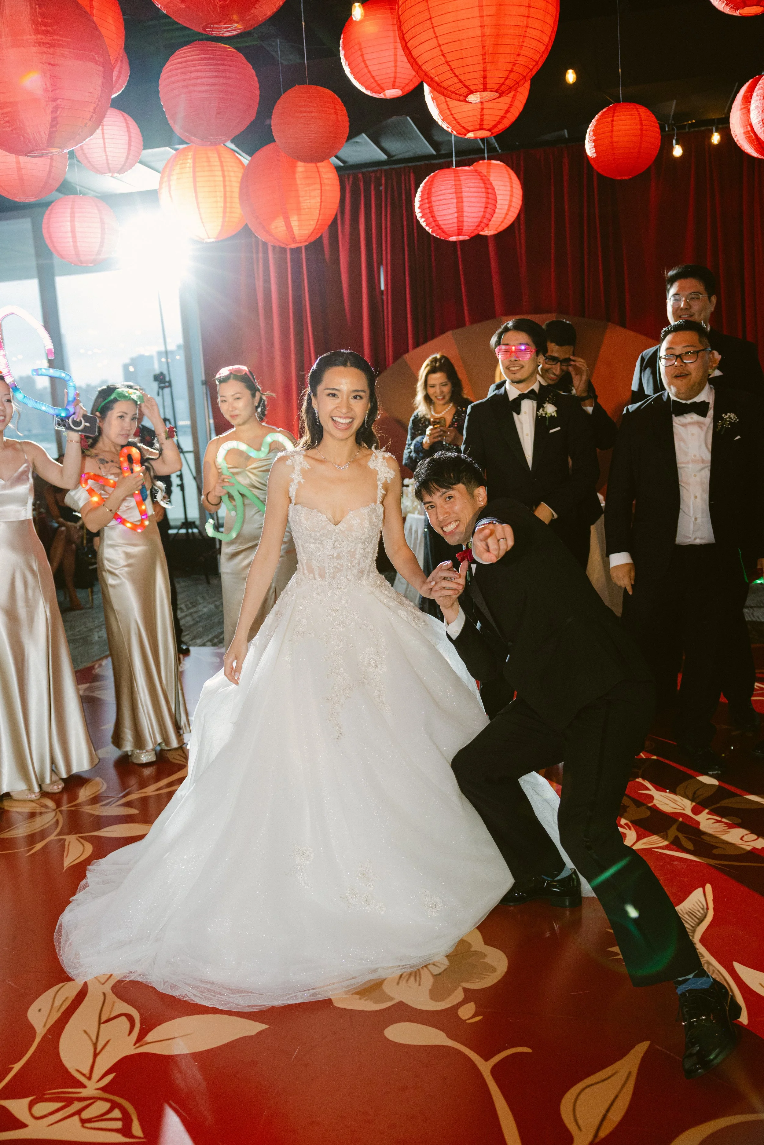 A bride in a white wedding gown and a groom in a tuxedo are dancing with friends and family at a wedding celebration. The background features red lanterns, curtains, and string lights, indicating an indoor festive event.