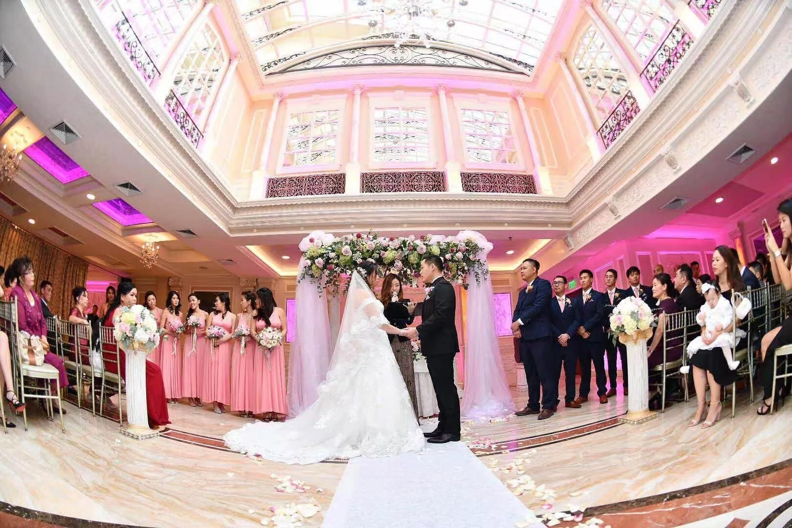A wedding ceremony taking place indoors with a bride and groom at the altar surrounded by bridesmaids and groomsmen, guests seated on chairs, lots of flowers, and pink lighting.
