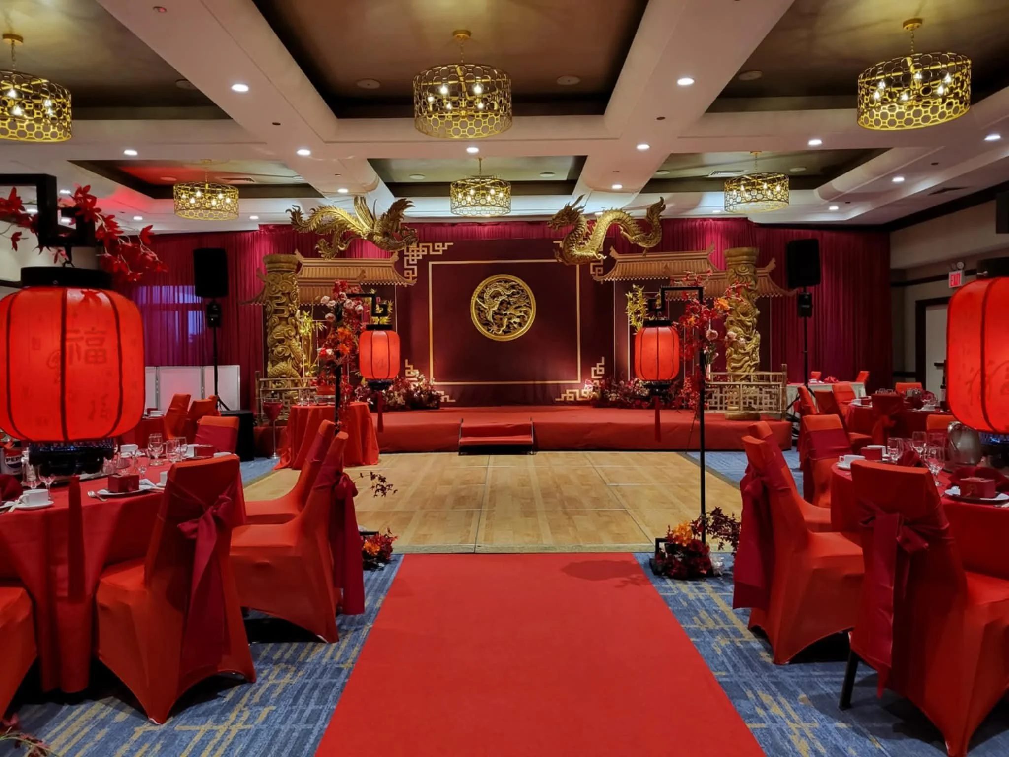 Traditional Asian-themed banquet hall decorated with red and gold, featuring a stage with golden dragons, red lanterns, and floral arrangements for an event or celebration.