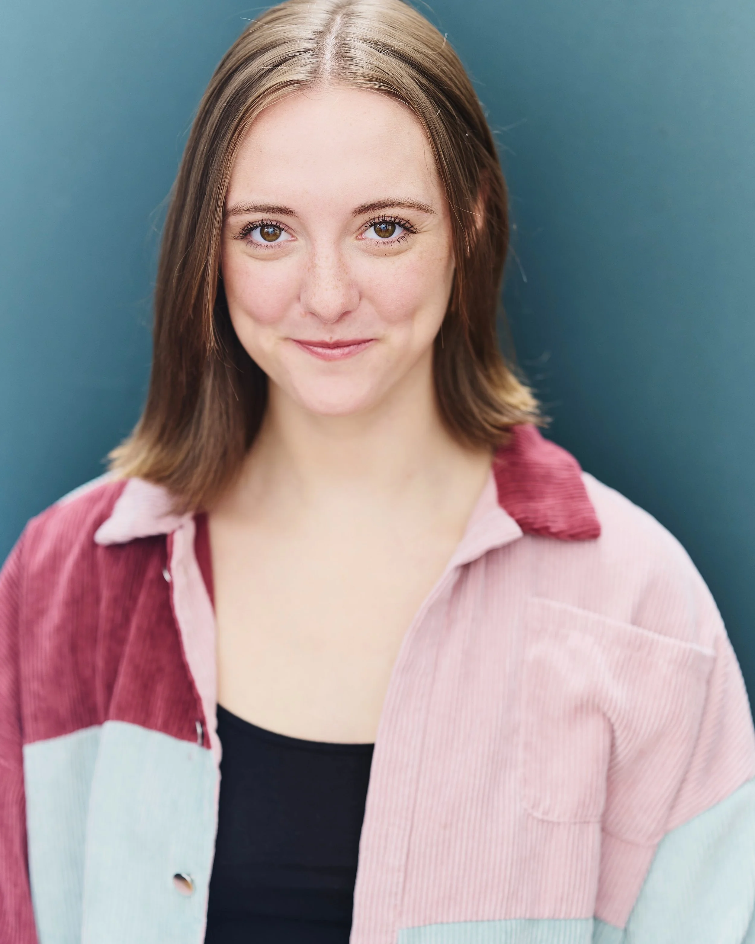 A young woman with brown hair, wearing a multicolored jacket and black shirt, smiling at the camera against a blue background.