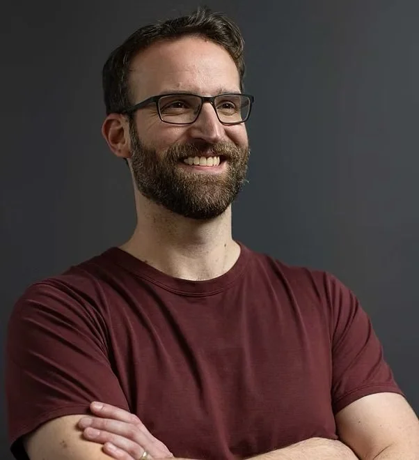 A smiling man with glasses and a beard, wearing a maroon T-shirt, standing with arms crossed against a dark gray background.