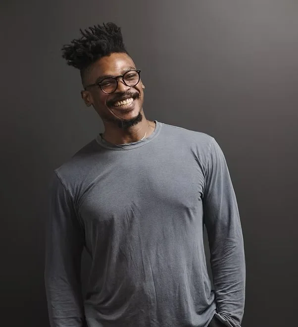A smiling man with glasses and a beard, wearing a gray long-sleeve shirt, standing against a gray background.