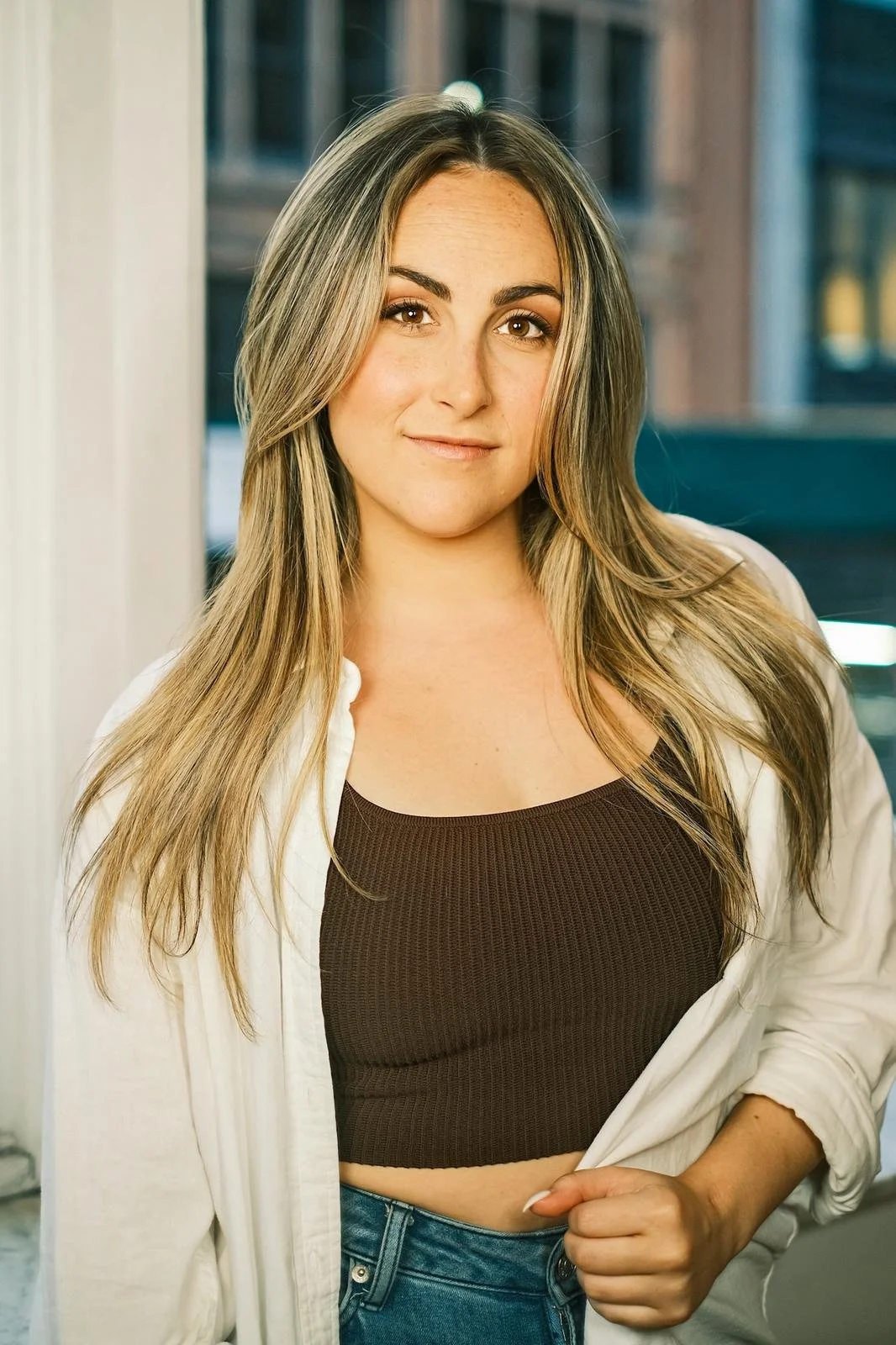 A young woman with long blonde hair, wearing a dark crop top and white shirt, standing indoors near a window with a cityscape background.