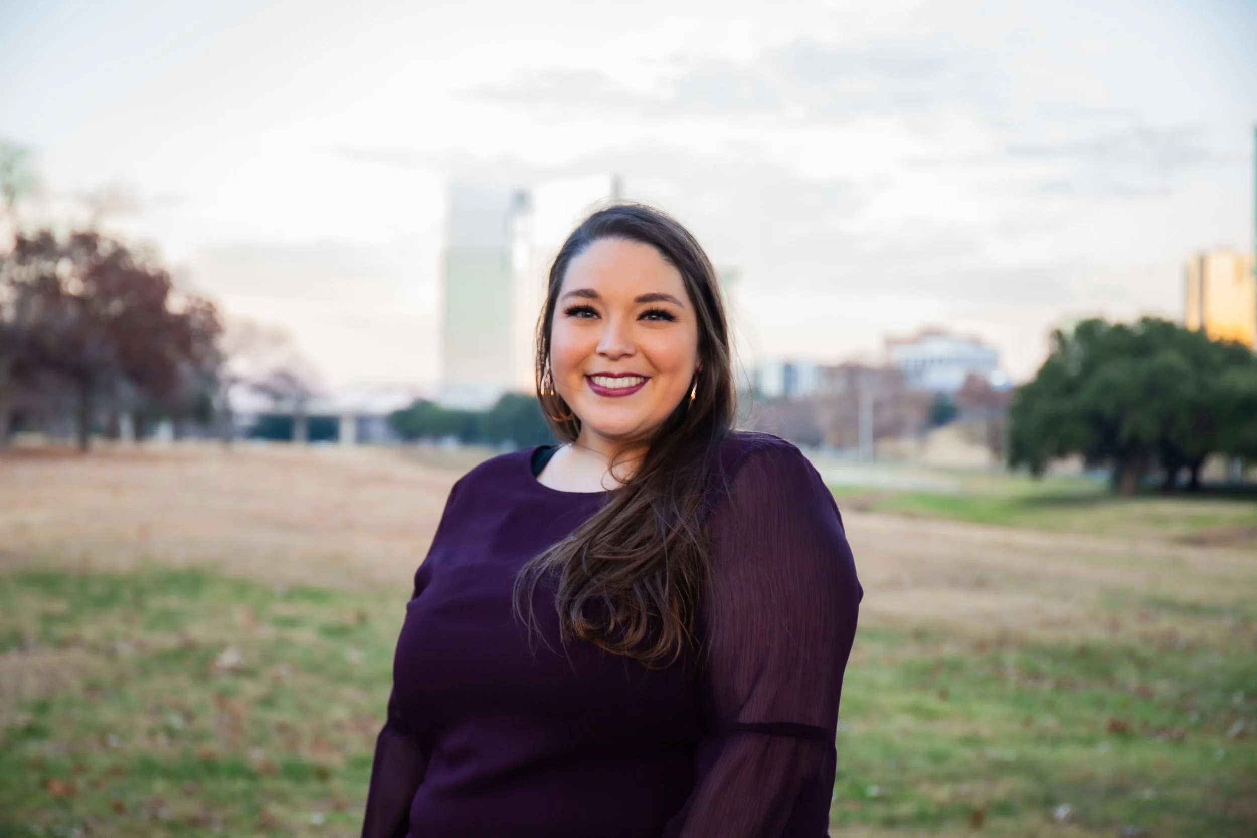 A young woman with long dark hair and earrings, smiling outdoors in a park with trees and buildings in the background.