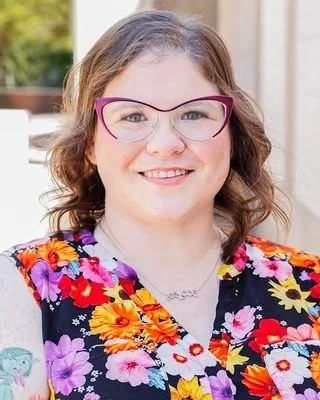 A woman with curly brown hair wearing glasses and a colorful floral top, smiling outdoors.