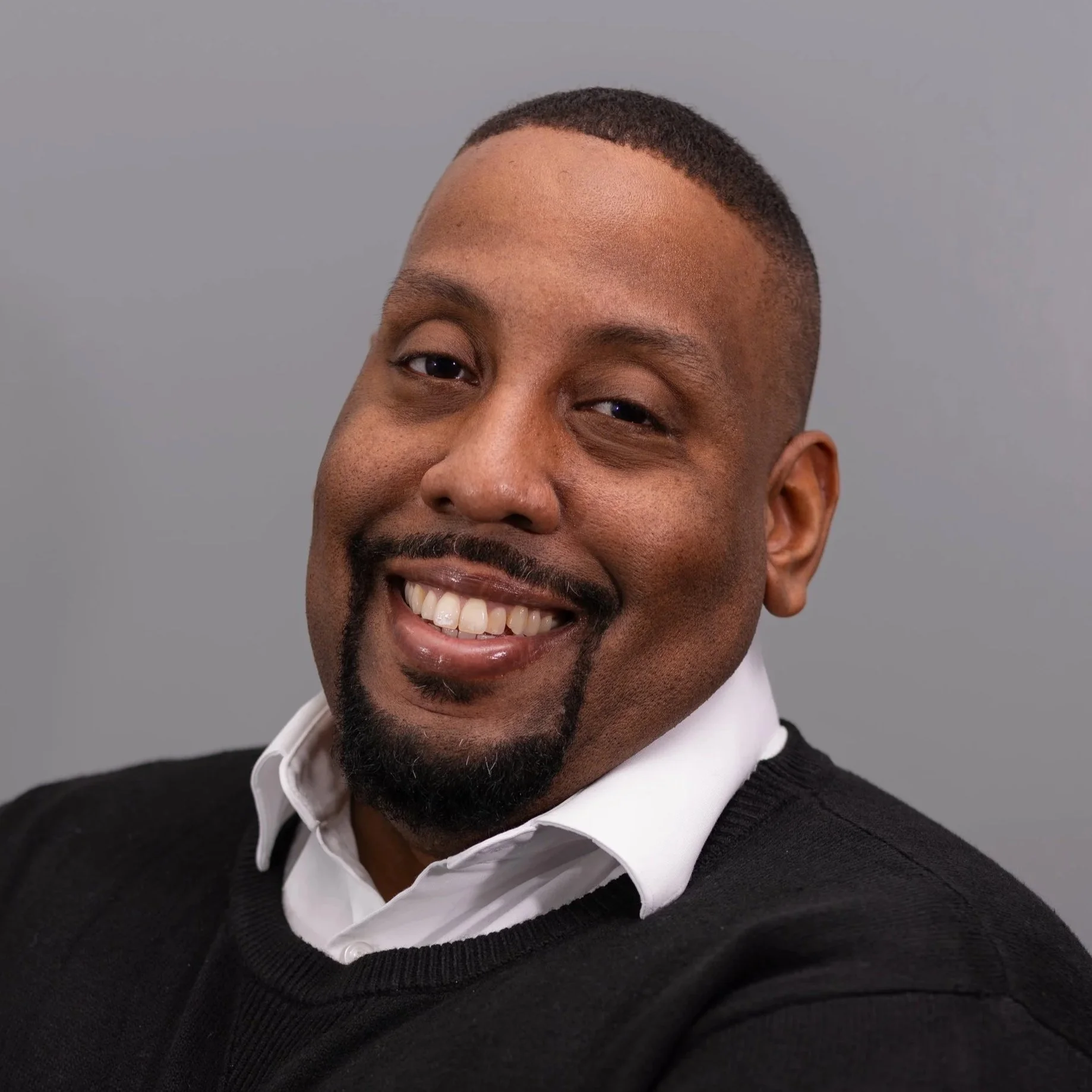 Headshot of a smiling African American man with short hair, a goatee, and a mustache, wearing a white shirt and black sweater, against a plain gray background.