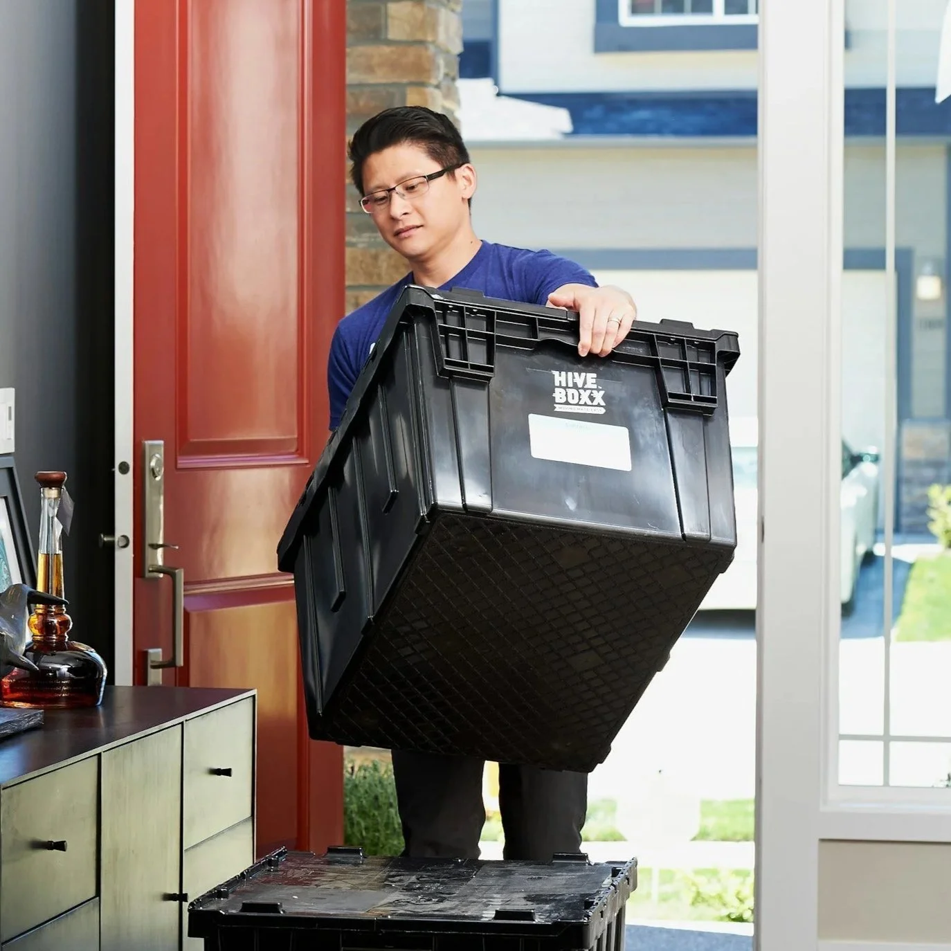 A man is lifting a large black plastic storage bin labeled 'HIVE BOXX' into or out of a house near an open door, with a smaller bin on the floor.