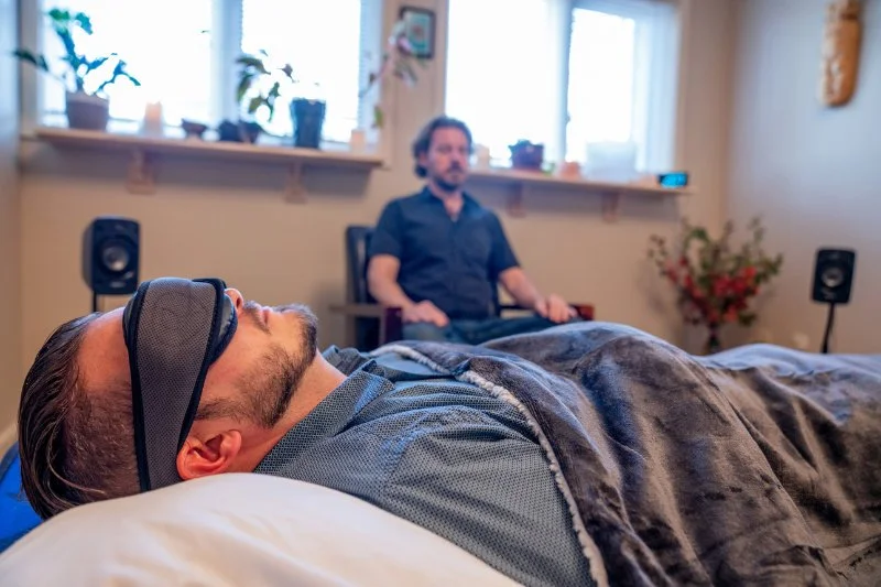 A man wearing a sleep mask is lying in bed while another man sits in a chair nearby, in a cozy room with large windows, plants, and speakers.