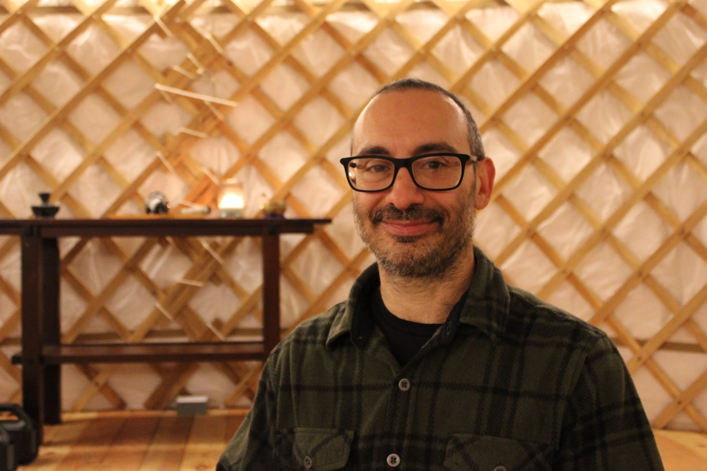 A man with glasses, beard, and short hair smiling in front of a lattice wall with a shelf and small objects on it.