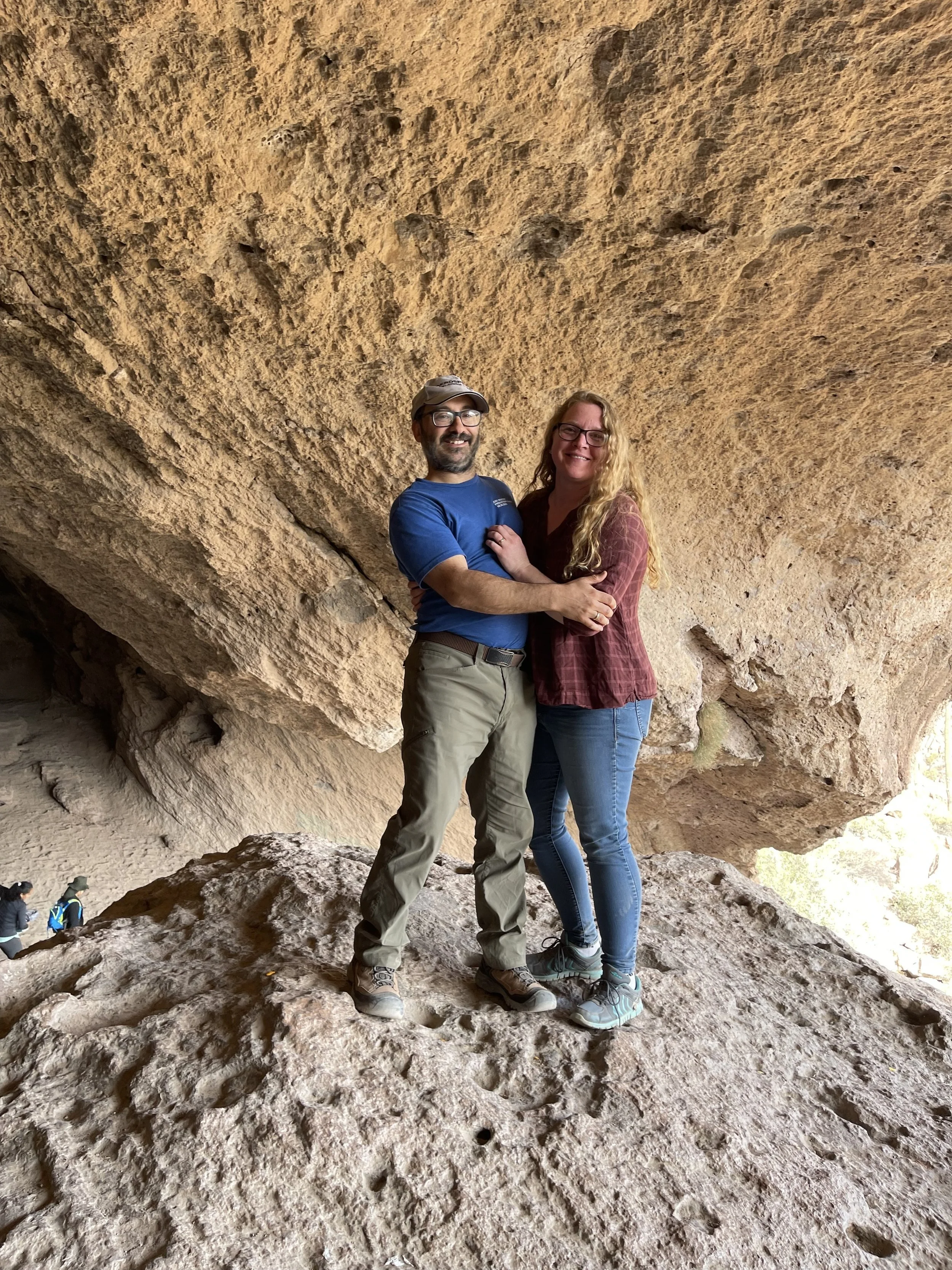 A man and woman standing close together inside a large, overhanging rock formation. The man wears a blue shirt, beige pants, glasses, and a cap. The woman wears a maroon plaid shirt and blue jeans. Both are smiling and looking at the camera.