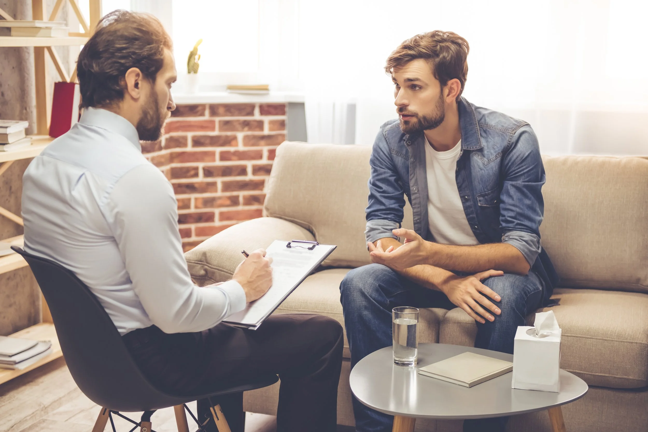A man with a beard and casual clothing talking to a therapist or counselor in an office.