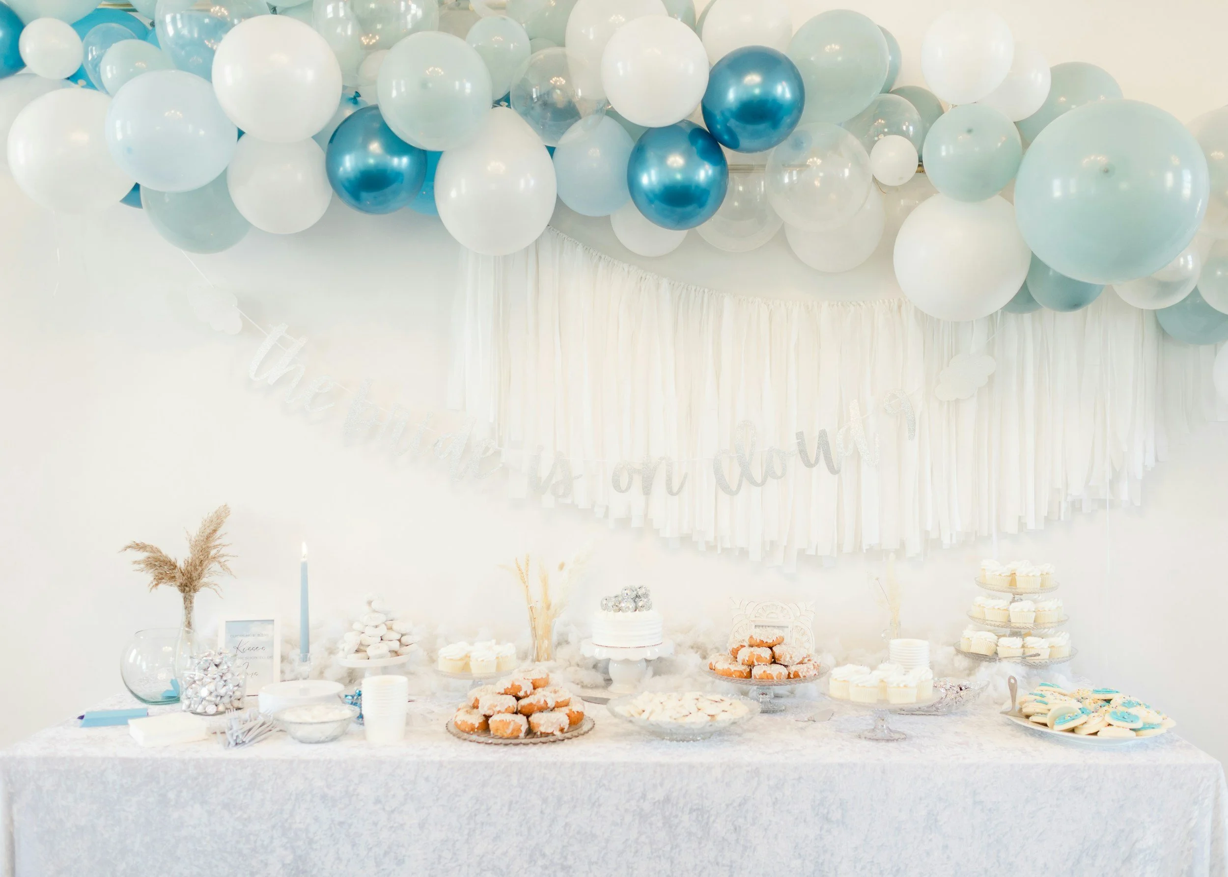 Dessert table decorated for a celebration with white and blue balloons overhead, a white tablecloth, and various pastries, cupcakes, and a cake. A banner in the background reads 'the party is on today'.