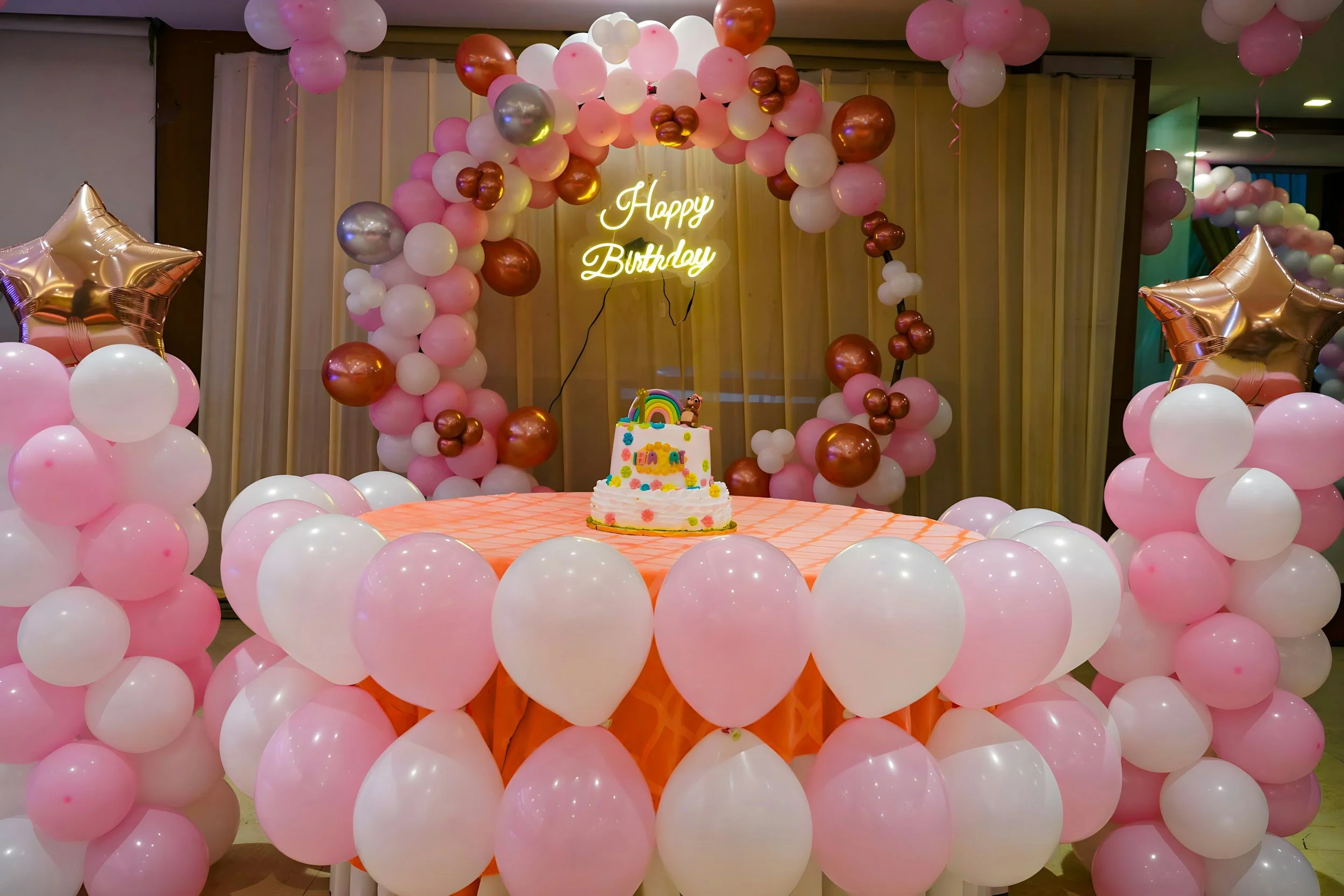Decorated birthday party table with pink and white balloons, a cake with rainbow and animal toppers, and a neon 'Happy Birthday' sign.