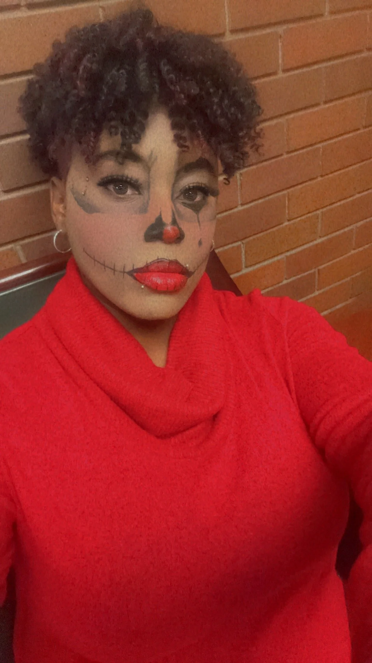 Woman with sugar skull face paint and red lipstick, wearing a red sweater, leaning against a brick wall.