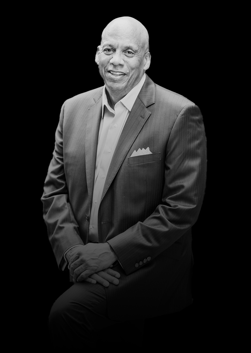 Black and white portrait of a smiling middle-aged man in a suit with a pocket square, sitting against a dark background.