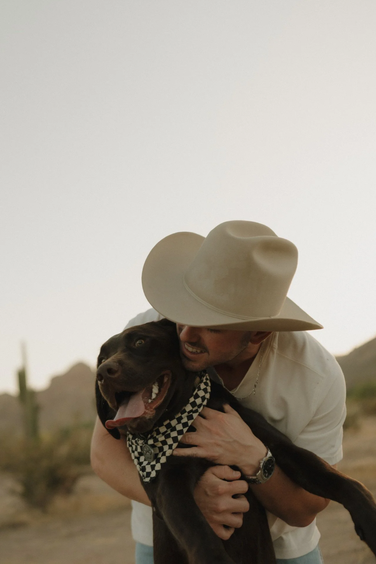 A man wearing a large beige cowboy hat hugging a happy chocolate Labrador retriever puppy with a black and white checkered bandana in a desert setting.