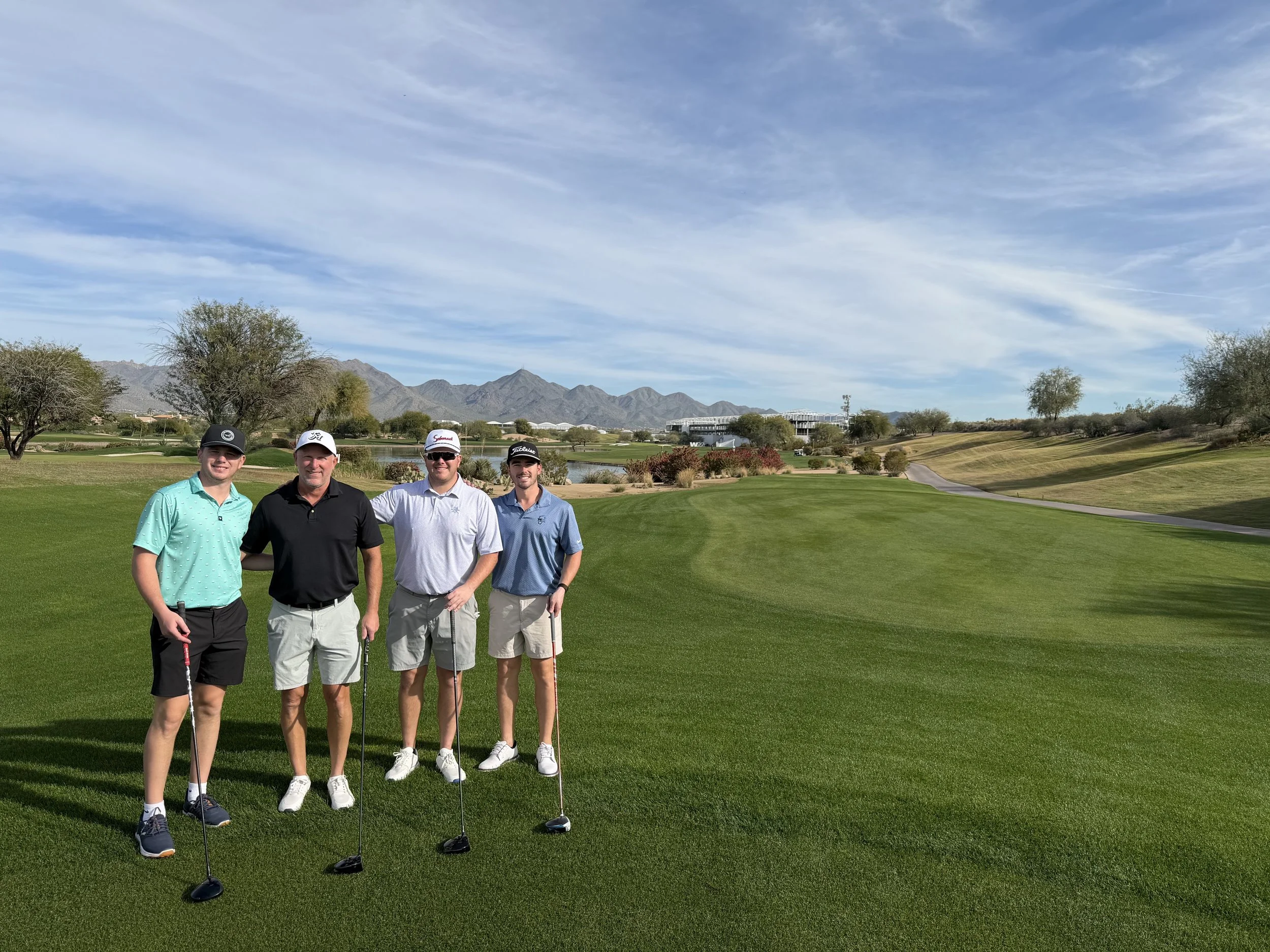 Four men on a golf course holding golf clubs, standing on the green with mountains and a clear sky in the background.