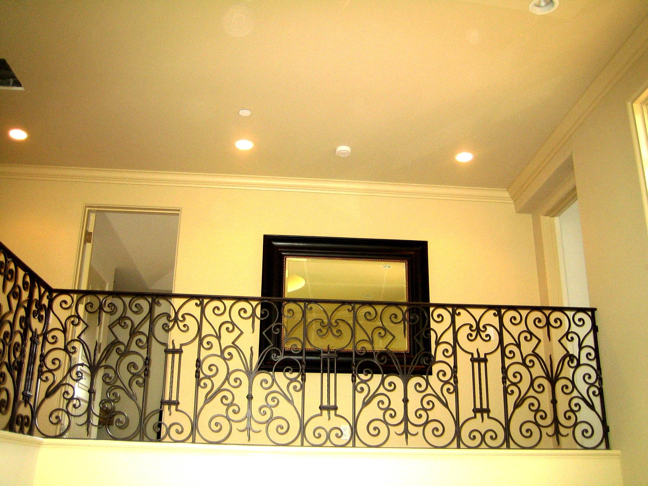 Interior view of a ceiling with four recessed lights, a mirror with a black frame on a cream wall, and an ornate black wrought iron railing.