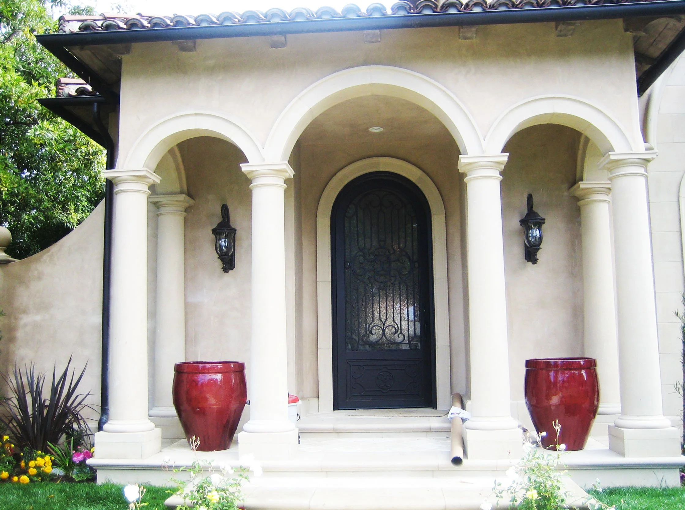 Front entrance of a house with arched columns, black front door with decorative metalwork, two black wall-mounted lanterns, large red planters, and steps leading to the door. Green lawn and trees in background.