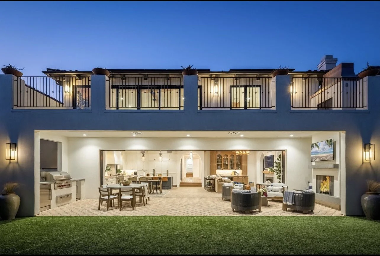 The rear view of a two-story modern house with a patio and balcony, illuminated at night. The ground level features an open living and dining area with furniture and a fireplace, visible through large sliding doors. The upper level has a balcony with