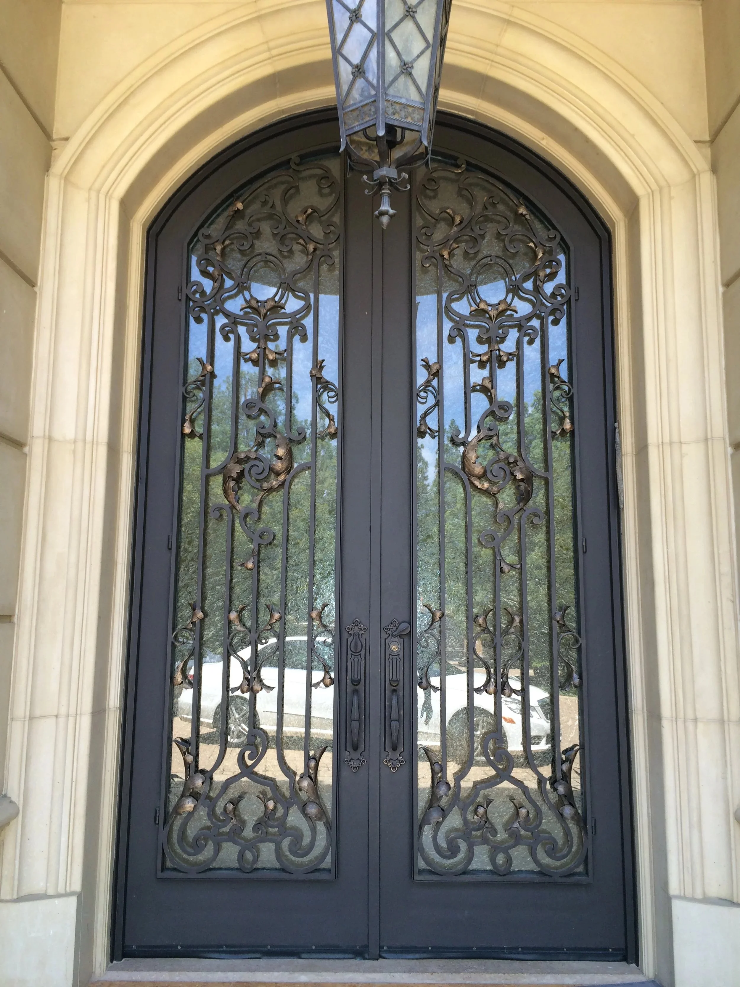 Double black wrought iron gate with ornate scrollwork design, set in a stone or concrete archway outside a building, with a reflection of trees and parked cars visible in the glass behind the gate.