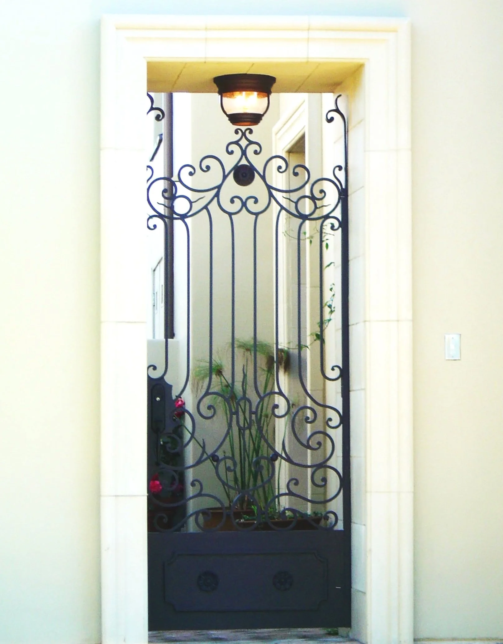 A decorative black wrought iron security door with intricate scrollwork pattern, behind which a potted plant is visible in an interior corridor. An overhead light fixture is mounted at the top of the doorframe.