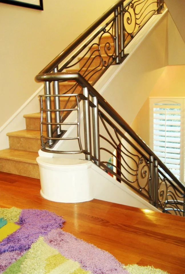 Interior view of a staircase with a wooden handrail and decorative black wrought iron balusters, beige carpeted stairs, and a wooden floor with a colorful rug.