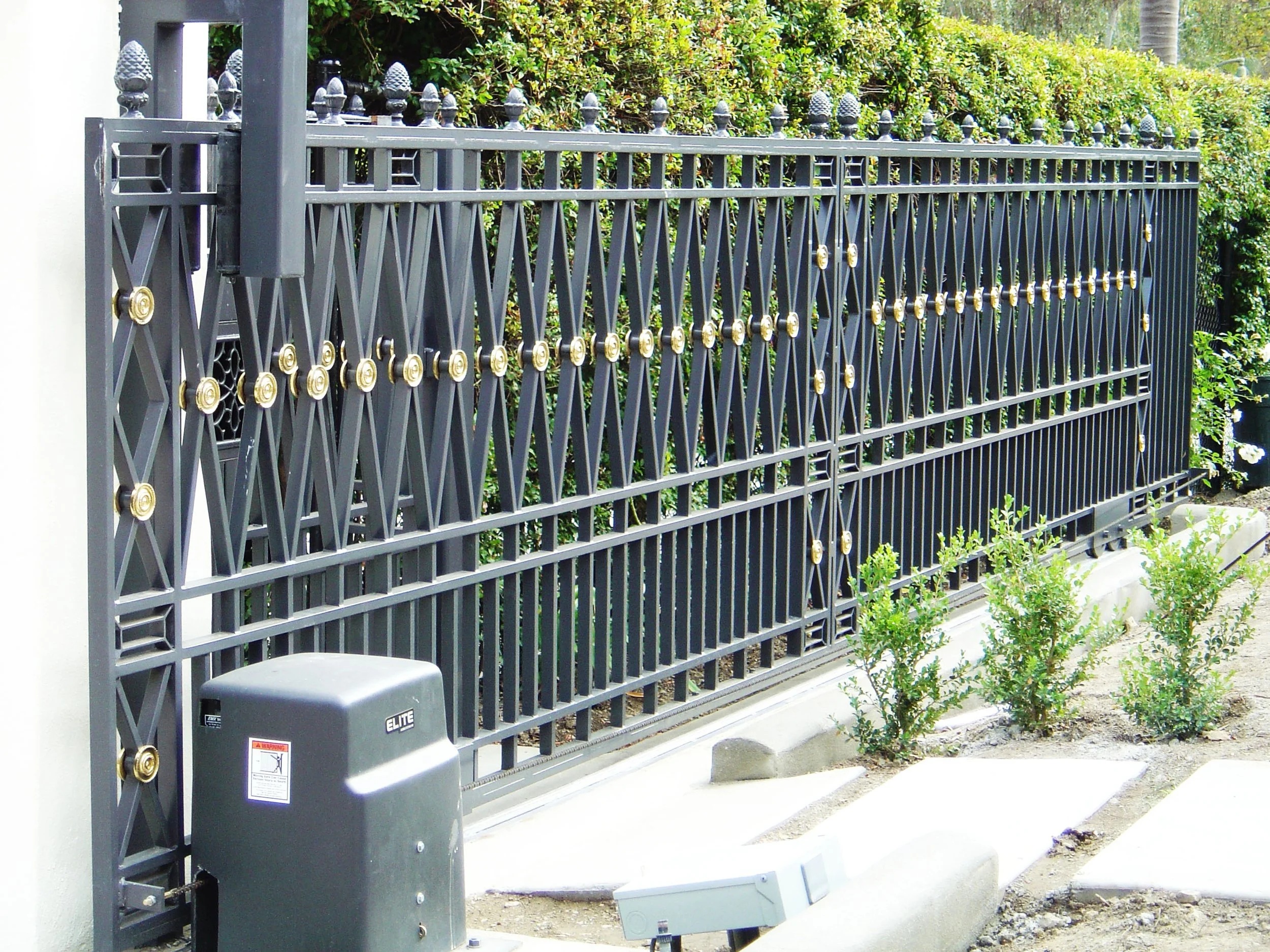 A black metal security gate with gold accents in front of greenery, with a gray control box on the left side and a row of small plants along the sidewalk.