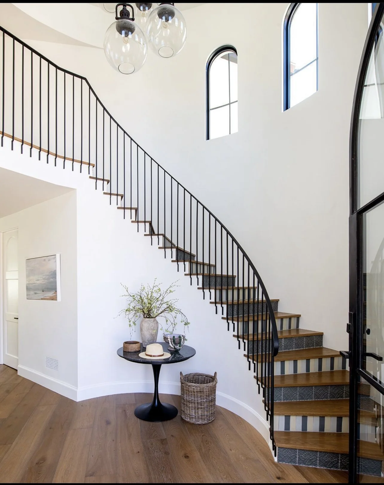Interior view of a modern staircase with black railing, wooden steps, and striped risers, next to a white wall with windows, a small round table with decorative items, and wooden flooring.
