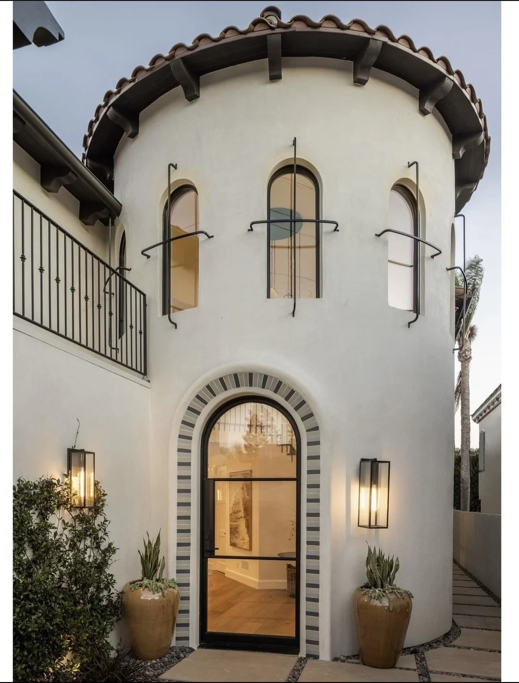 Front entrance of a Mediterranean-style house with a rounded tower, arched windows, potted plants, and wall-mounted lanterns.