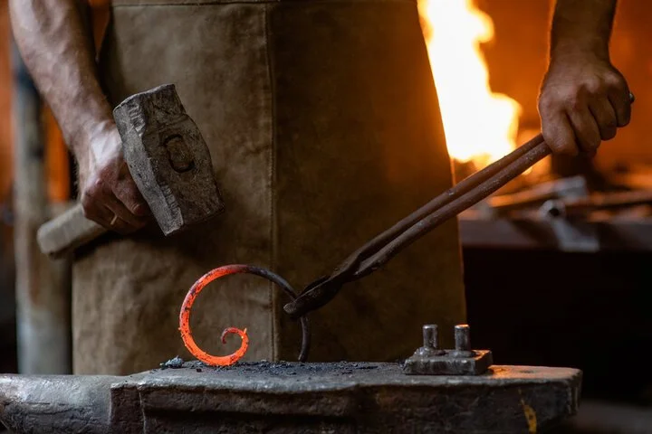 Blacksmith working with heated metal, holding a hammer and tongs in a workshop with a forge fire in the background.