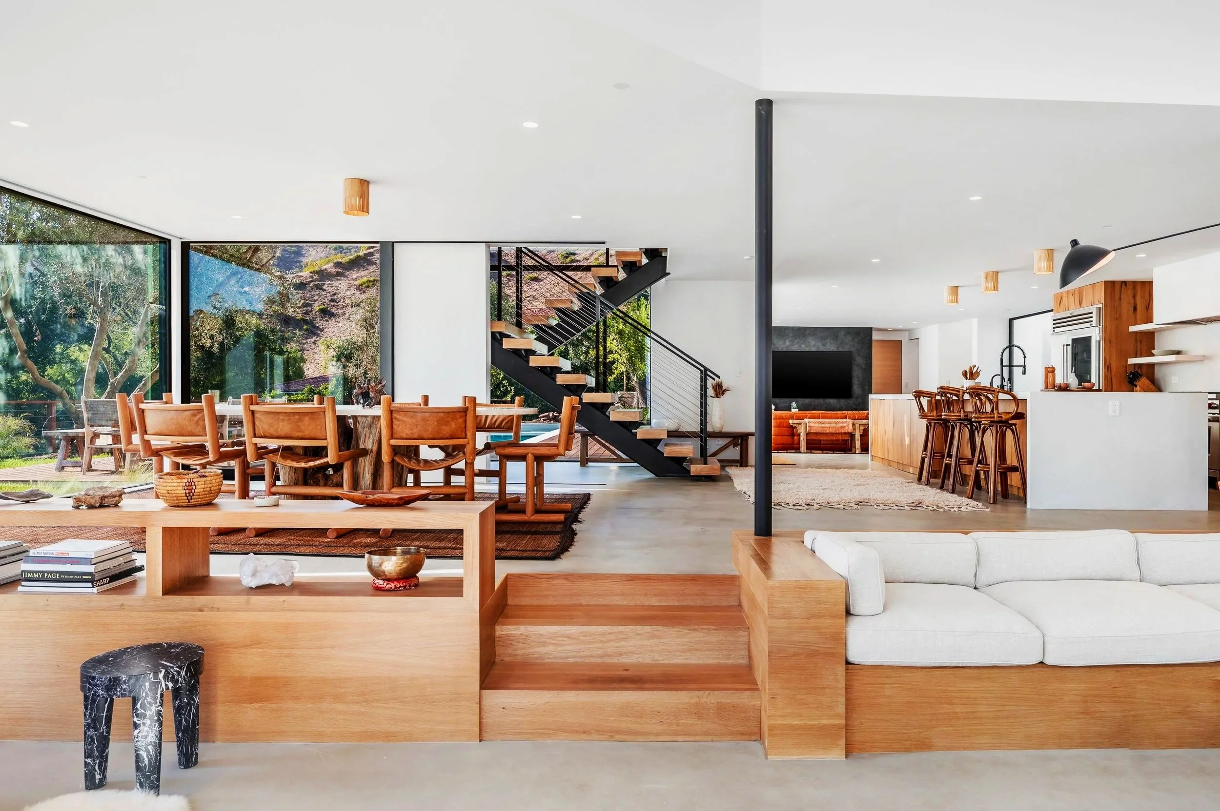 Open-concept living and dining room with wooden furniture, large windows, modern staircase, and kitchen area in the background.