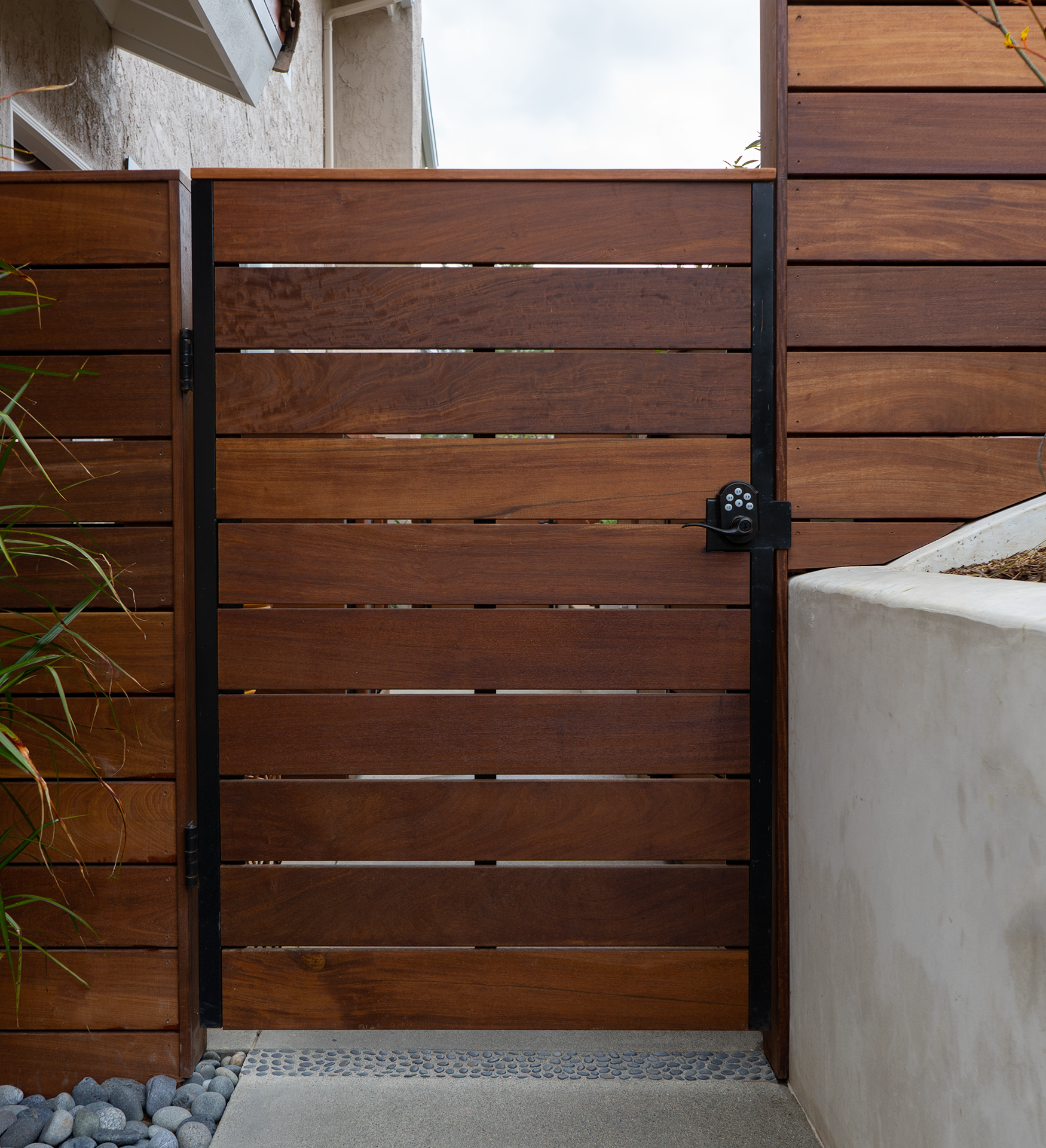 A wooden gate with black hardware and a combination lock, situated between a wooden fence and a concrete wall, with pebbles and a doormat on the ground.