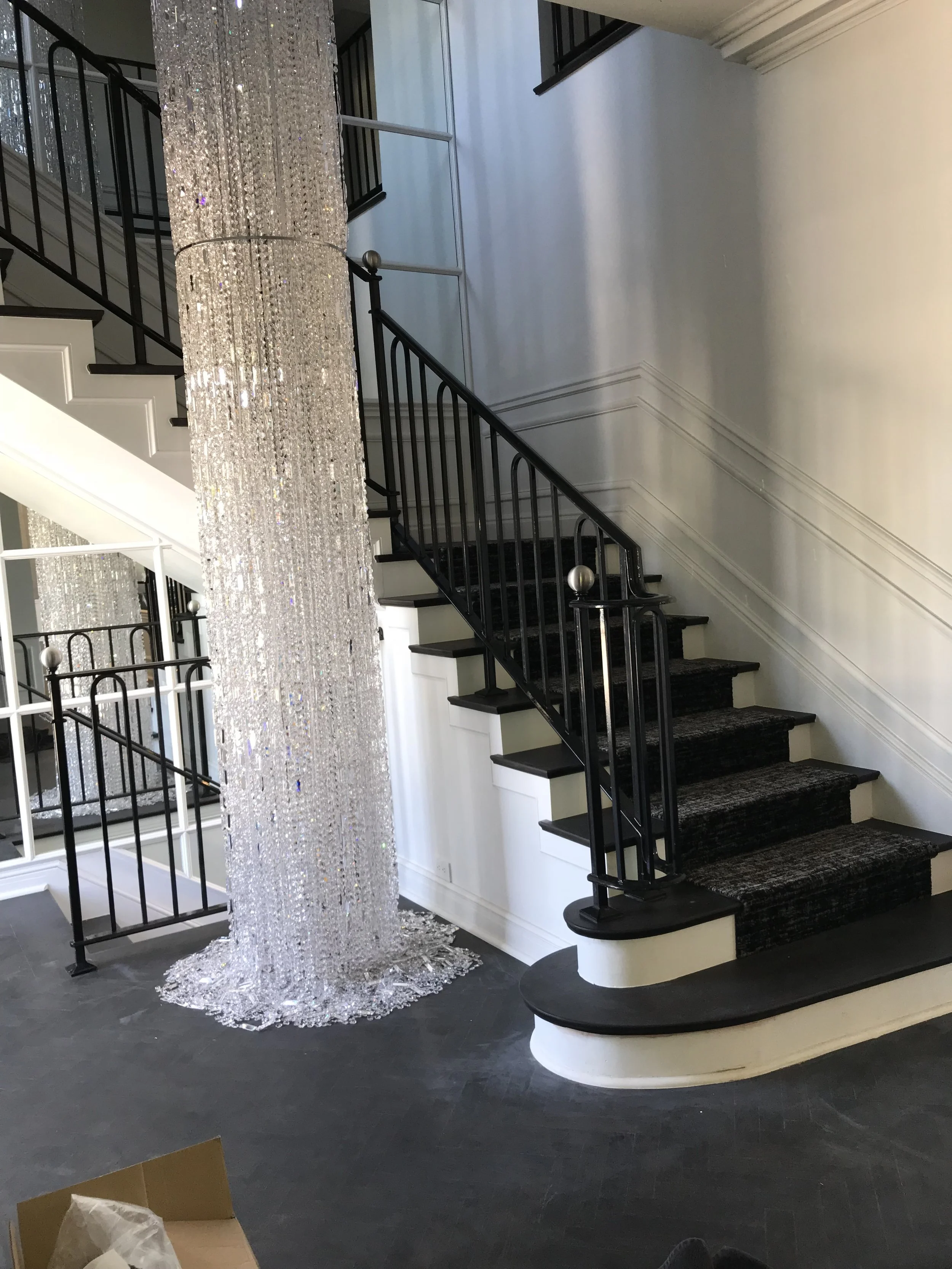 A grand staircase with black railing and marble steps is next to a large hanging chandelier made of numerous crystals, in a well-lit, elegant interior space.