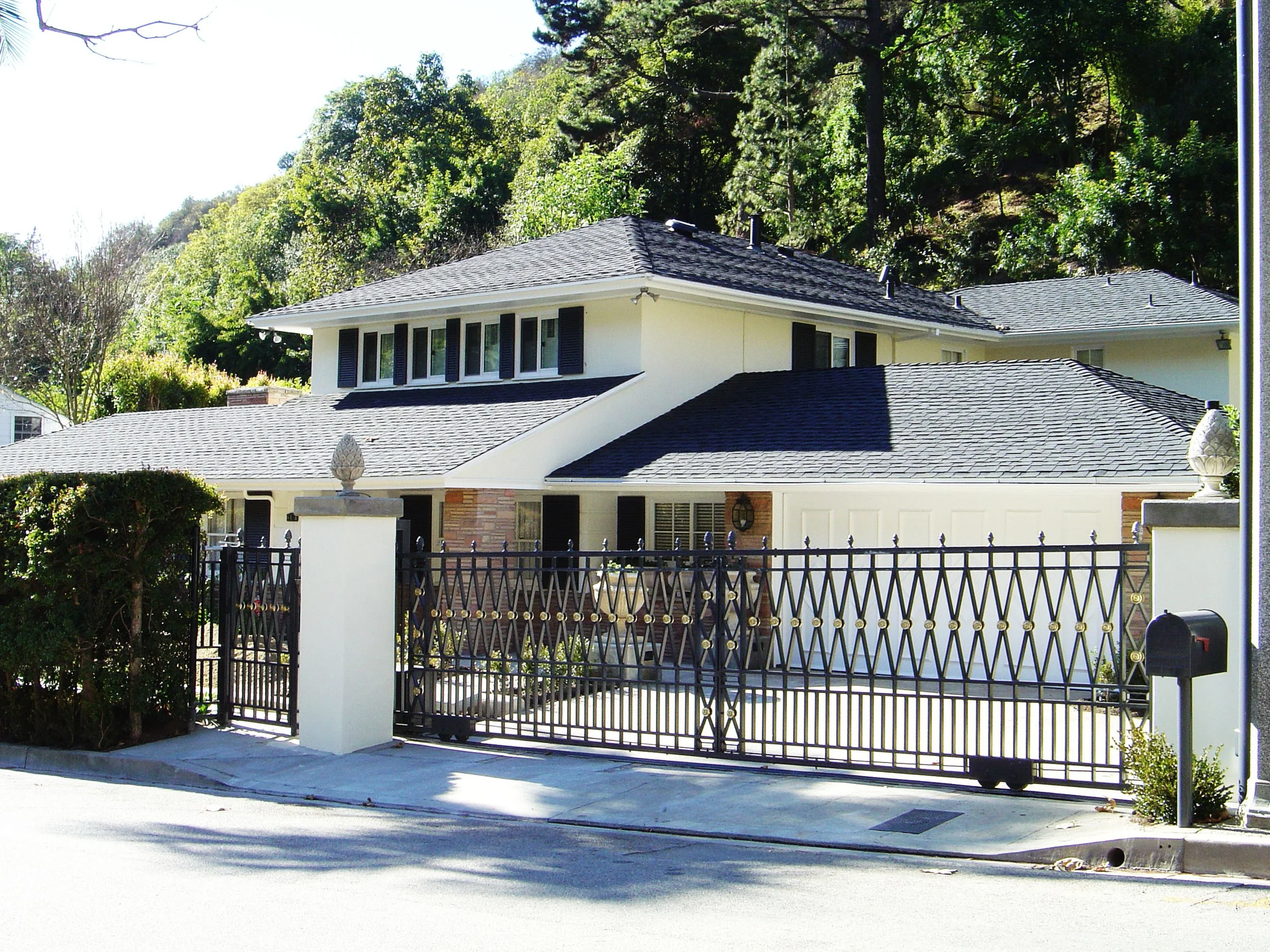 A two-story white house with a dark shingle roof, black shutters on the windows, and a driveway enclosed by a black wrought iron gate with stone pillars, situated in a green, hilly area.