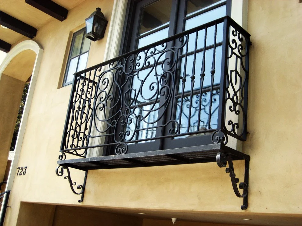 Decorative black metal balcony with scrolling design on a beige stucco building wall.
