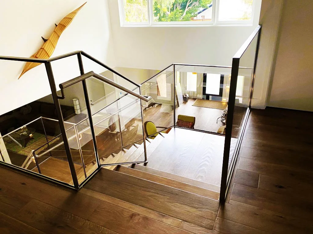 View of a staircase from above, with glass railings, leading down to a living area with wooden floors and a large window.