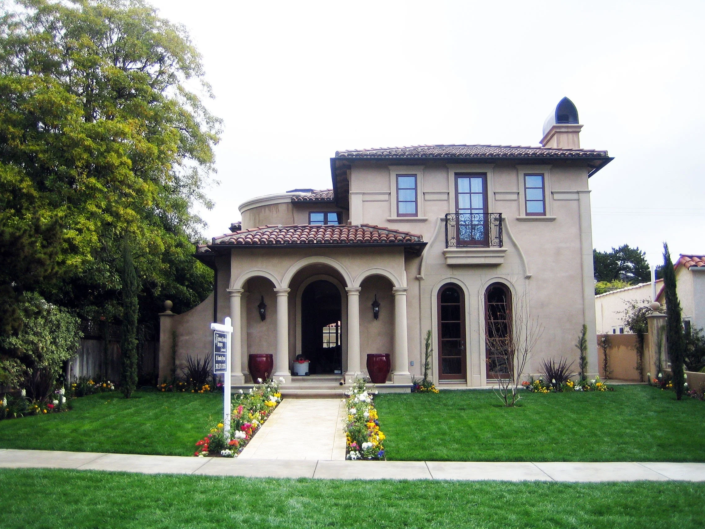 A two-story house with beige stucco walls, a red tile roof, and a small balcony in the front. The front yard has a well-manicured lawn, flower beds, and a paved walkway leading to the entrance.