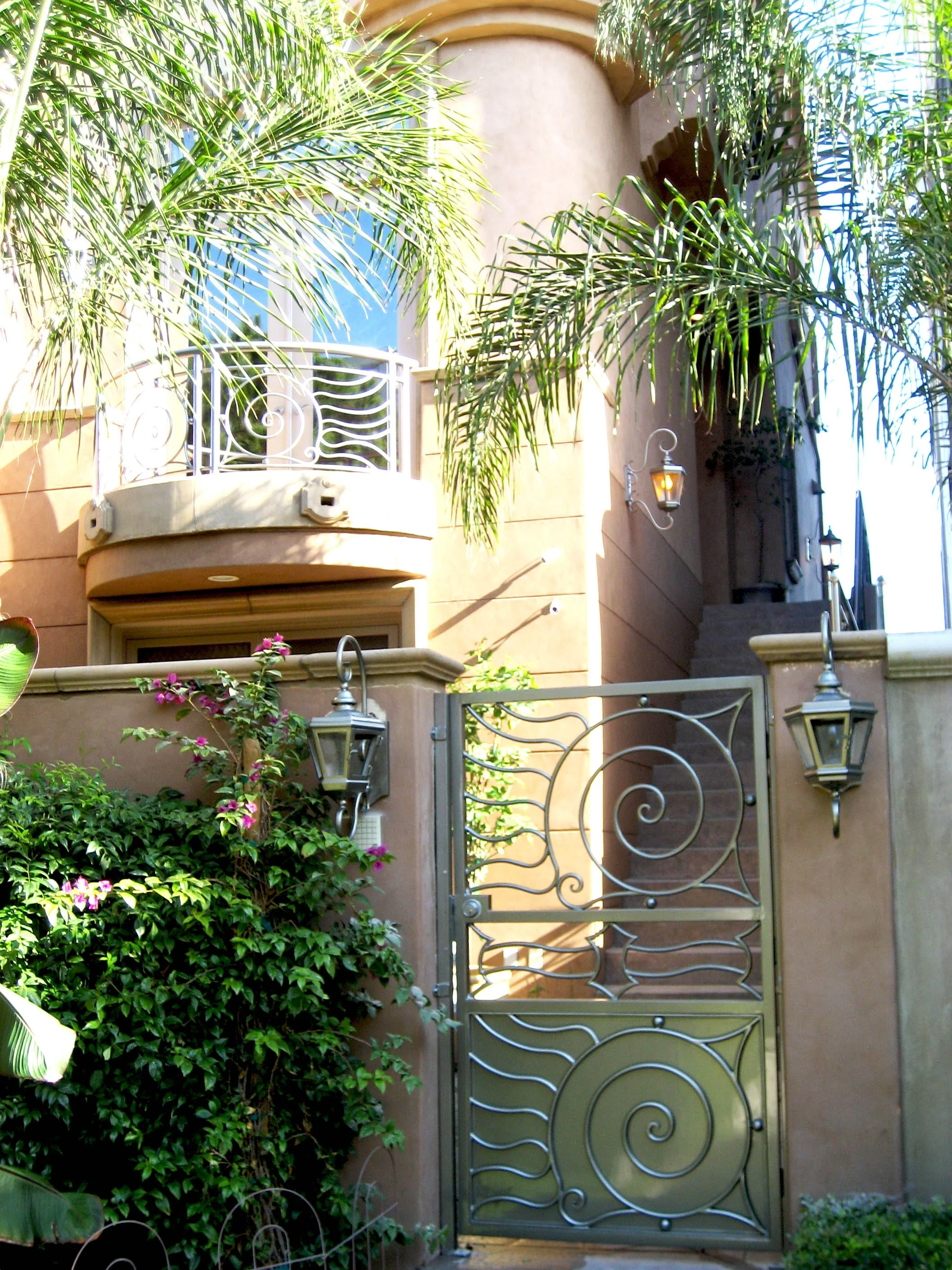 A gated entrance to a house with decorative metalwork, surrounded by lush green plants and palm trees, with exterior lighting fixtures.