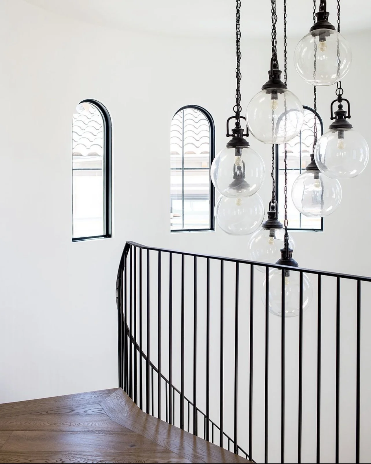 Modern staircase with black metal railing, wooden step, white wall with three tall, narrow arched windows, and a hanging chandelier with multiple glass globes.