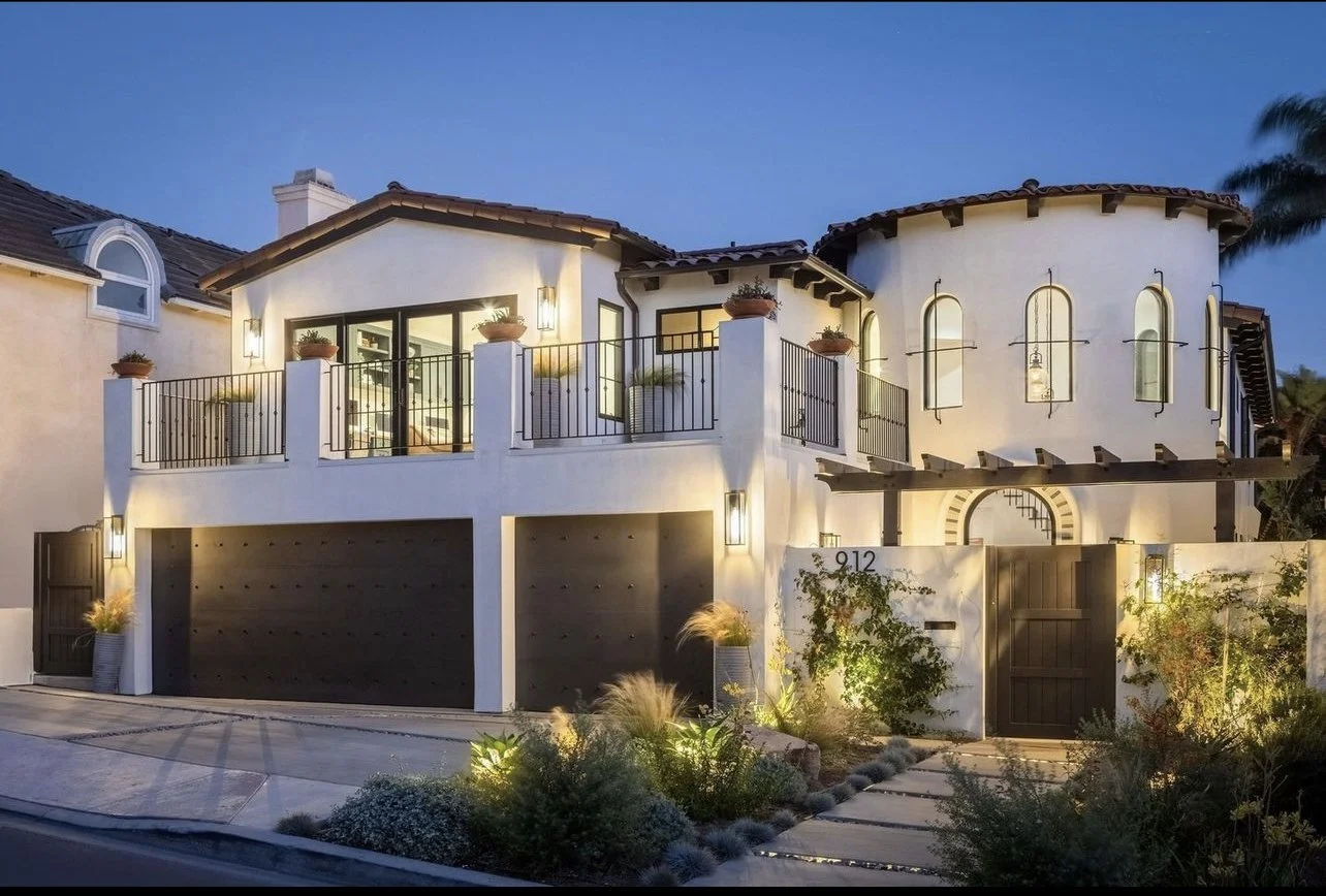 A modern two-story house with a white stucco exterior, black accents, and a curved tower section. It has a spacious balcony with potted plants and outdoor lighting, and a landscaped front yard with various plants and a pathway.