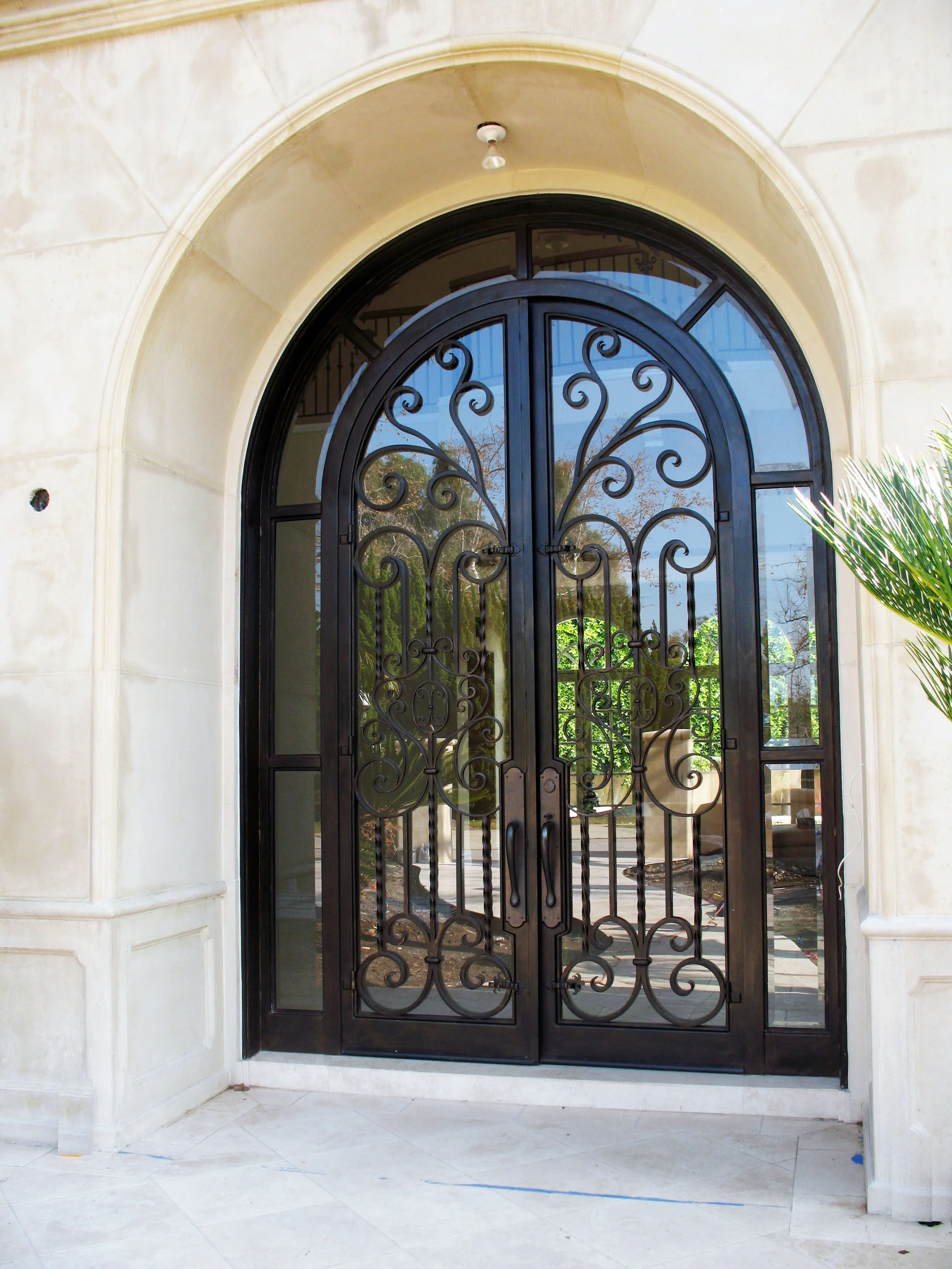 Black wrought iron double door with decorative scrollwork, set in a cream stone colonial-style archway, with a view of indoor stairs and greenery reflected in the glass.