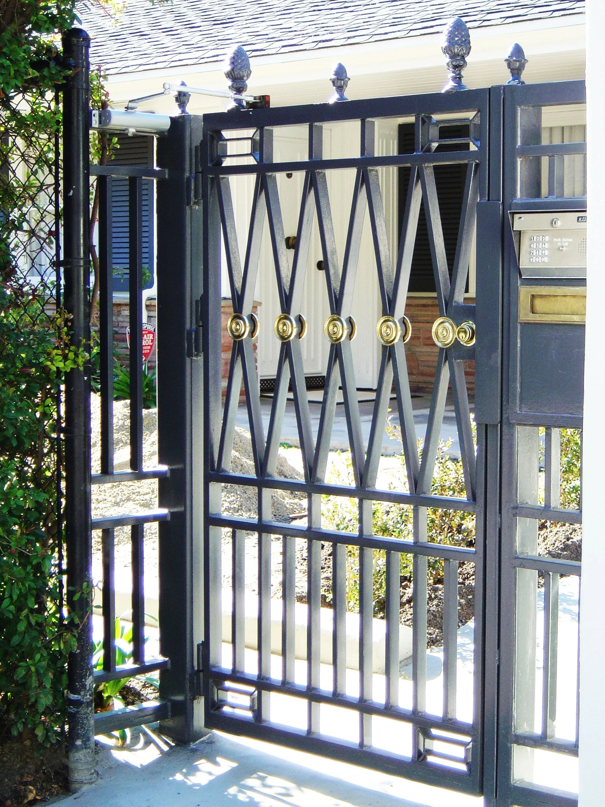 Black decorative wrought iron gate with gold accents, mail slot, and keypad, opening to a home with shrubs and a porch